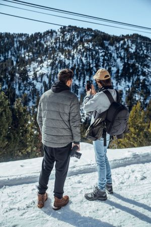 Men with smartphone and camera on hill between trees in winter