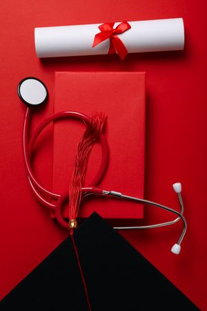 Graduate hat with diploma and stethoscope, on red background.