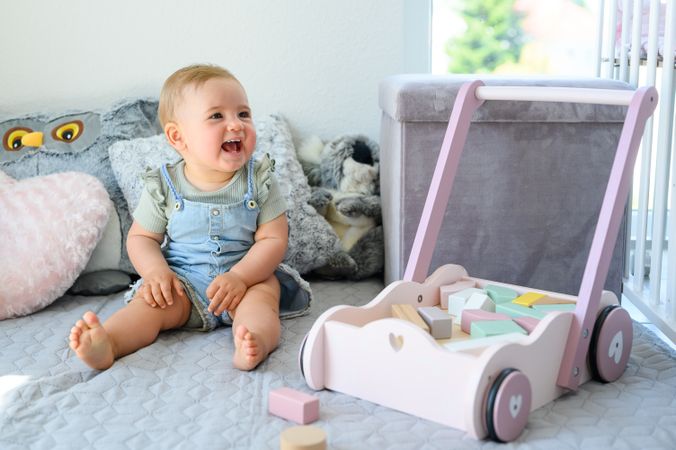 Happy little baby sitting near wooden blocks in cart on bed