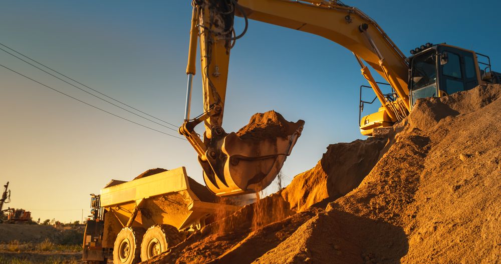 Construction Site On Sunny Evening: Industrial Excavator Loading Sand Into A Truck. The Process Of Building New Apartment Block. Workers Operating Heavy Machinery To Complete Real Estate Project.