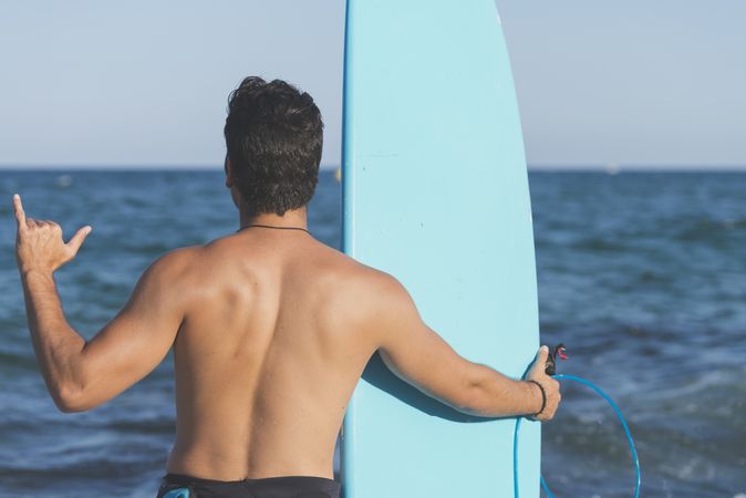 Male surfer with blue board standing in front of the water making Shaka sign with hand