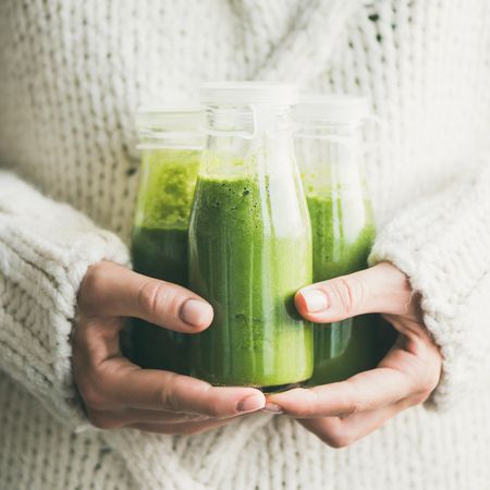 Woman holding three bottles of green smoothie wearing cozy sweater, square crop